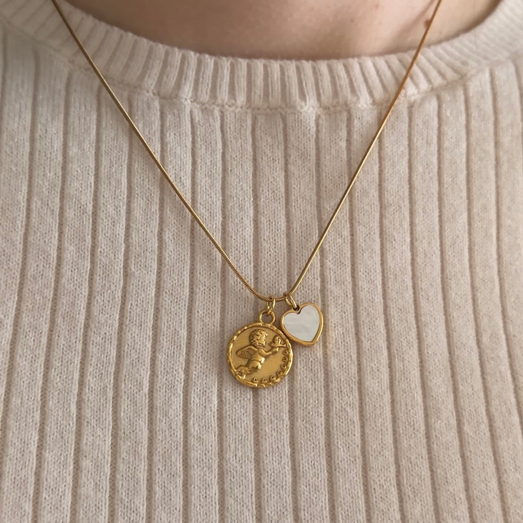A close-up of a person wearing a gold necklace with two pendants: a round coin-style charm with a Cherub and a small white heart-shaped charm, styled over a white ribbed sweater. 