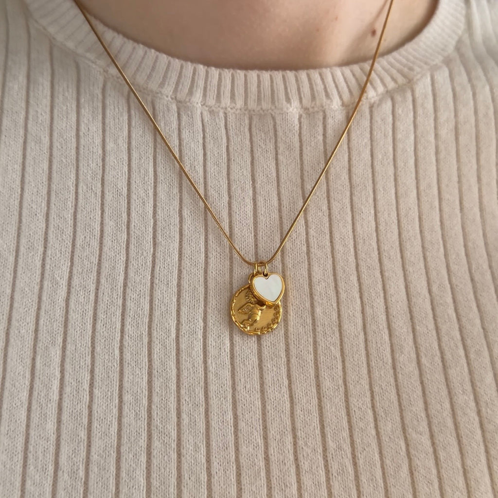 A close-up of a person wearing a gold necklace with two pendants: a round coin-style charm with a Cherub and a small white heart-shaped charm, styled over a white ribbed sweater. 