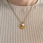 A close-up of a person wearing a gold necklace with two pendants: a round coin-style charm with a Cherub and a small white heart-shaped charm, styled over a white ribbed sweater. 
