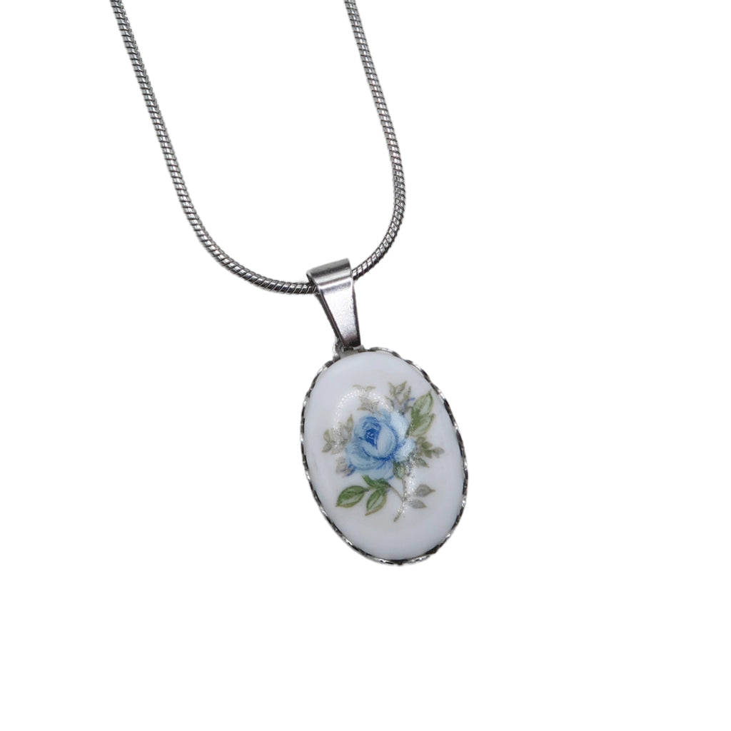 An oval pendant necklace featuring a white cabochon with a delicate blue rose and green leaves, set in a silver bezel on a silver snake chain from the side