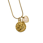 A gold-tone chain necklace with two charms: a textured, embossed round coin pendant with a cherub and a small heart-shaped charm with a smooth, milky-white inlay, laid against a plain white background.