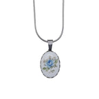 An oval pendant necklace featuring a white cabochon with a delicate blue rose and green leaves, set in a silver bezel on a silver snake chain.